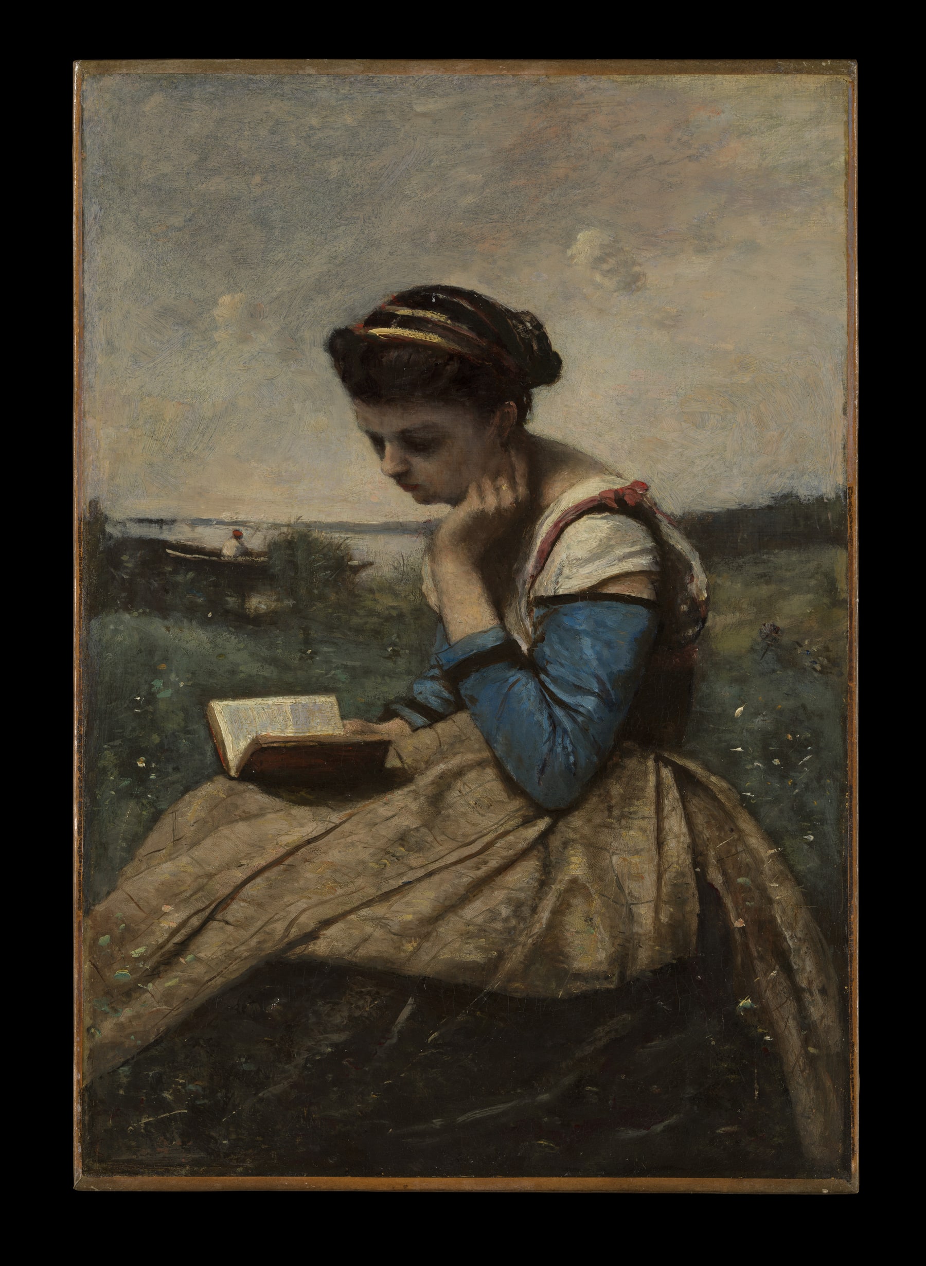 A woman in a blue blouse reads an open book in her lap beside the water.
